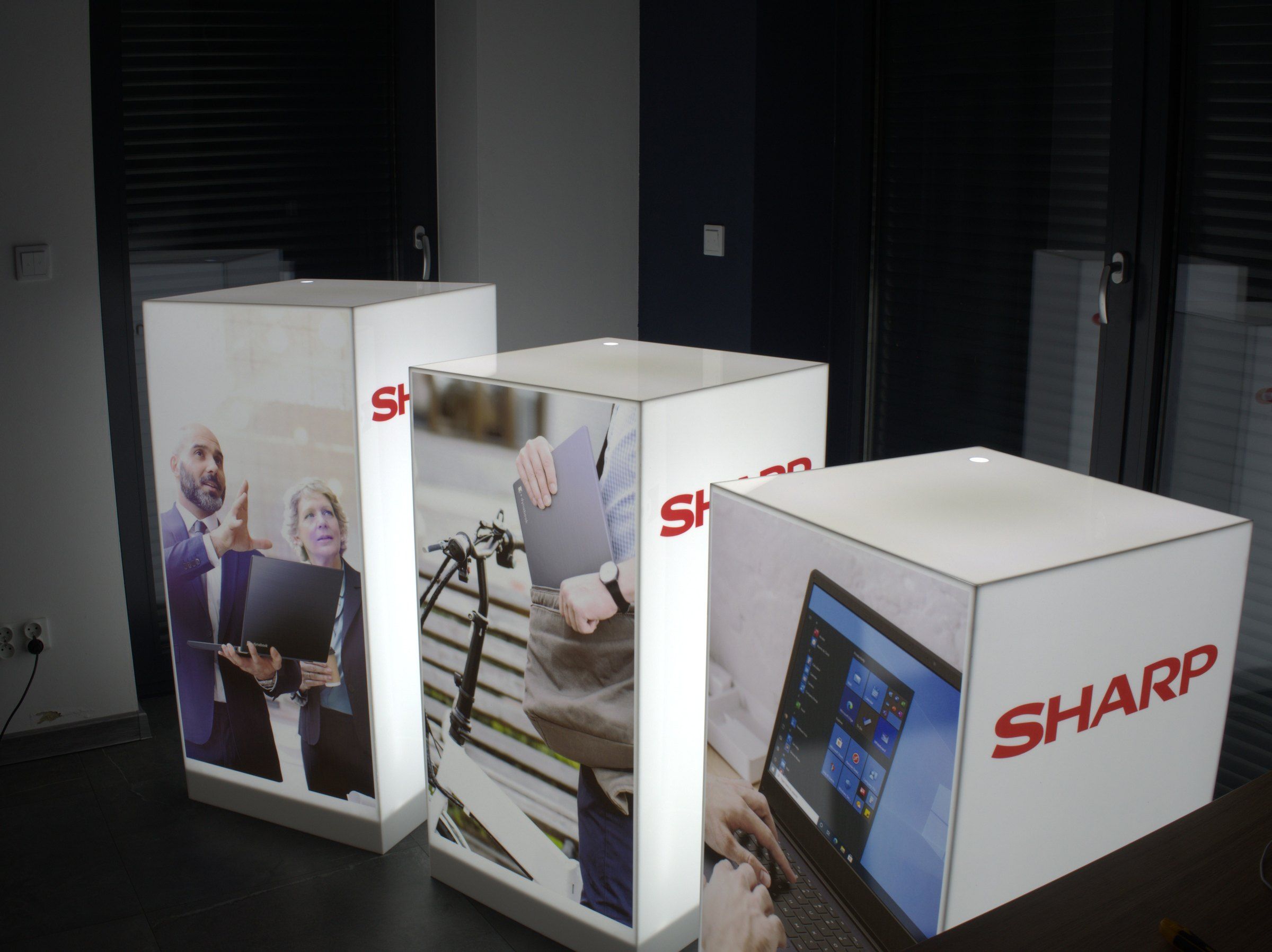 Trzy podświetlane kubiki ekspozycyjne różnej wysokości z grafikami i logotypami Sharp i Dynabook w showroomie